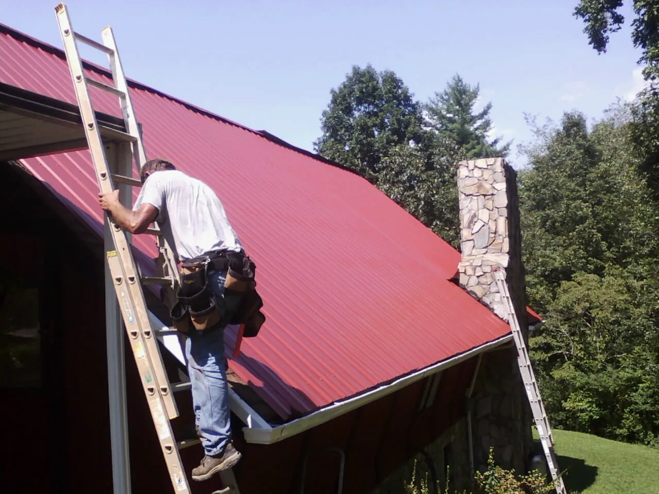 Roofer on a ladder beside a red metal roof and stone chimney — Soffit & Fascia Repair by Roofing Brothers in Bluff City